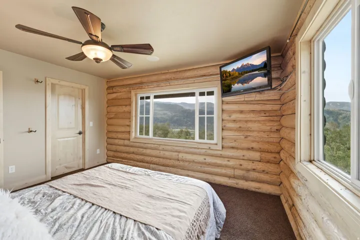 Natural wood-log wall, large windows, ceiling fan with integrated lighting, and a mounted flat-screen television