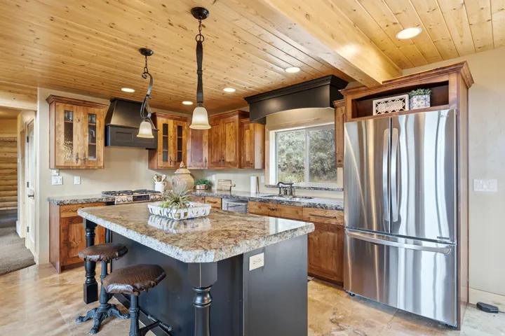 Wood-paneled ceiling with recessed lighting, stainless steel French door refrigerator, black range hood, wood cabinetry, and a kitchen island with stone countertop