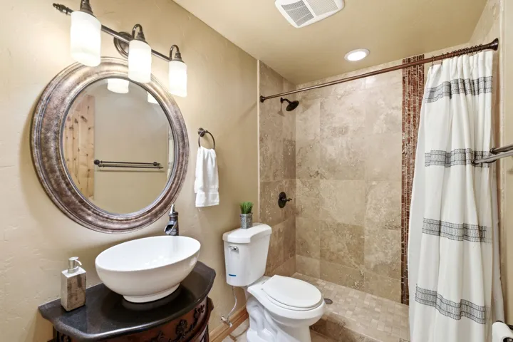 Vessel sink with dark countertop, ornate vanity, round framed mirror, three-light vanity fixture, and tiled shower with accent band