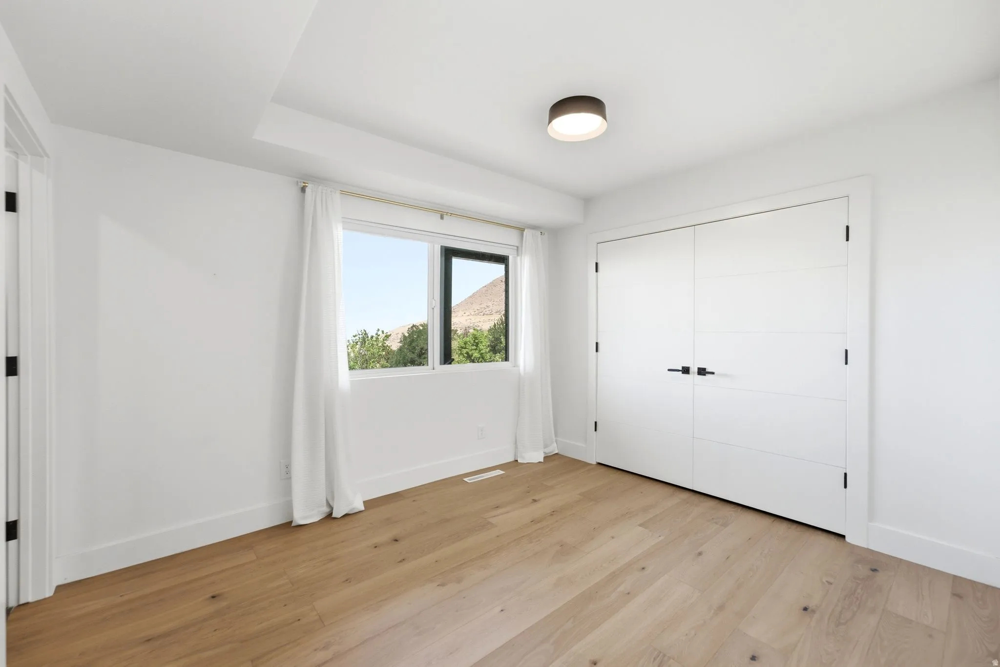 Light wood-finish flooring, white walls, modern flush mount lighting, a large window, and double closet doors with matte black hardware