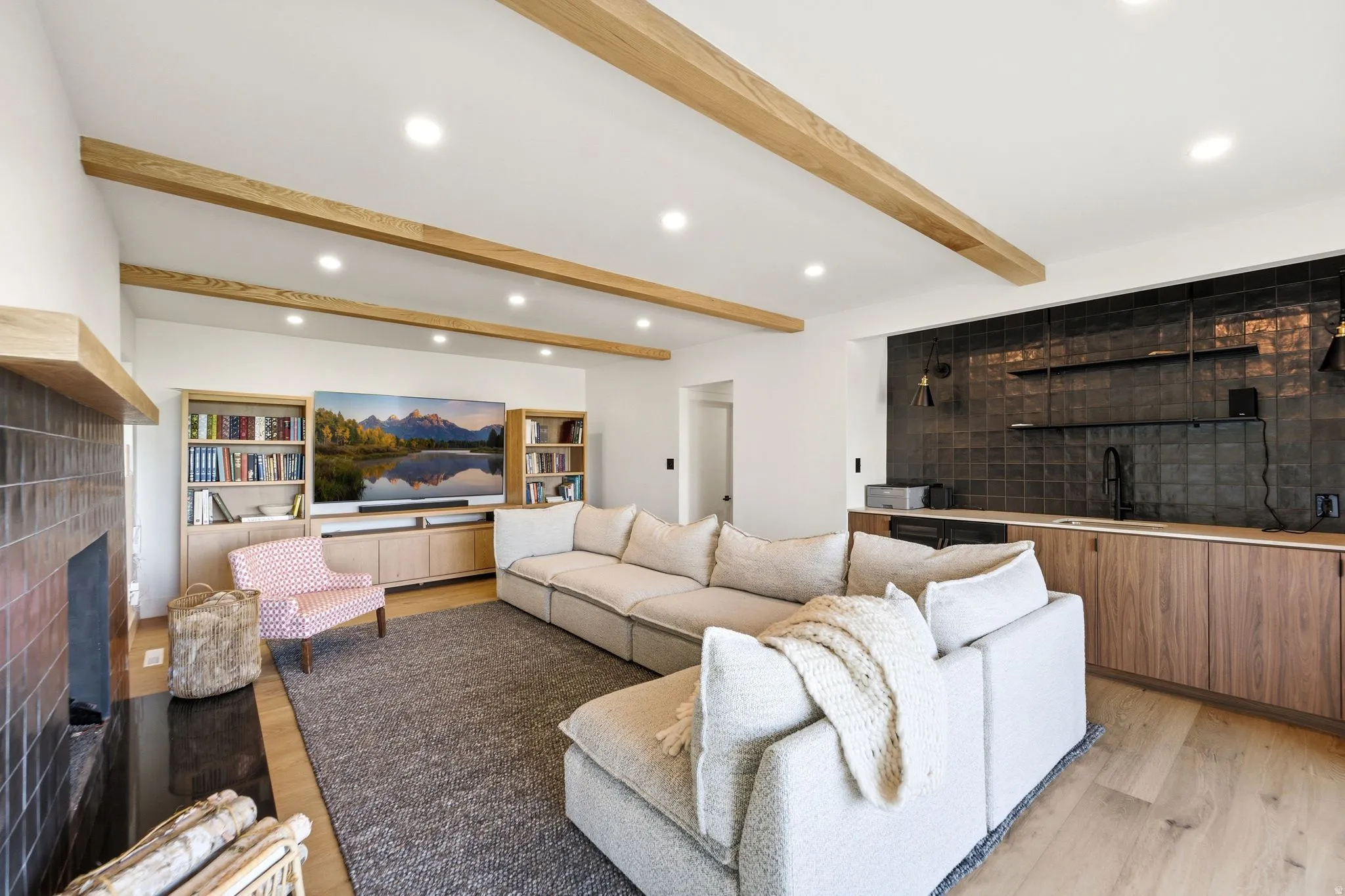 Vaulted ceiling with exposed wood beams, wood-finish flooring, built-in media cabinetry with integrated shelving, fireplace with tiled surround, and wet bar featuring tiled backsplash and wood cabinetry