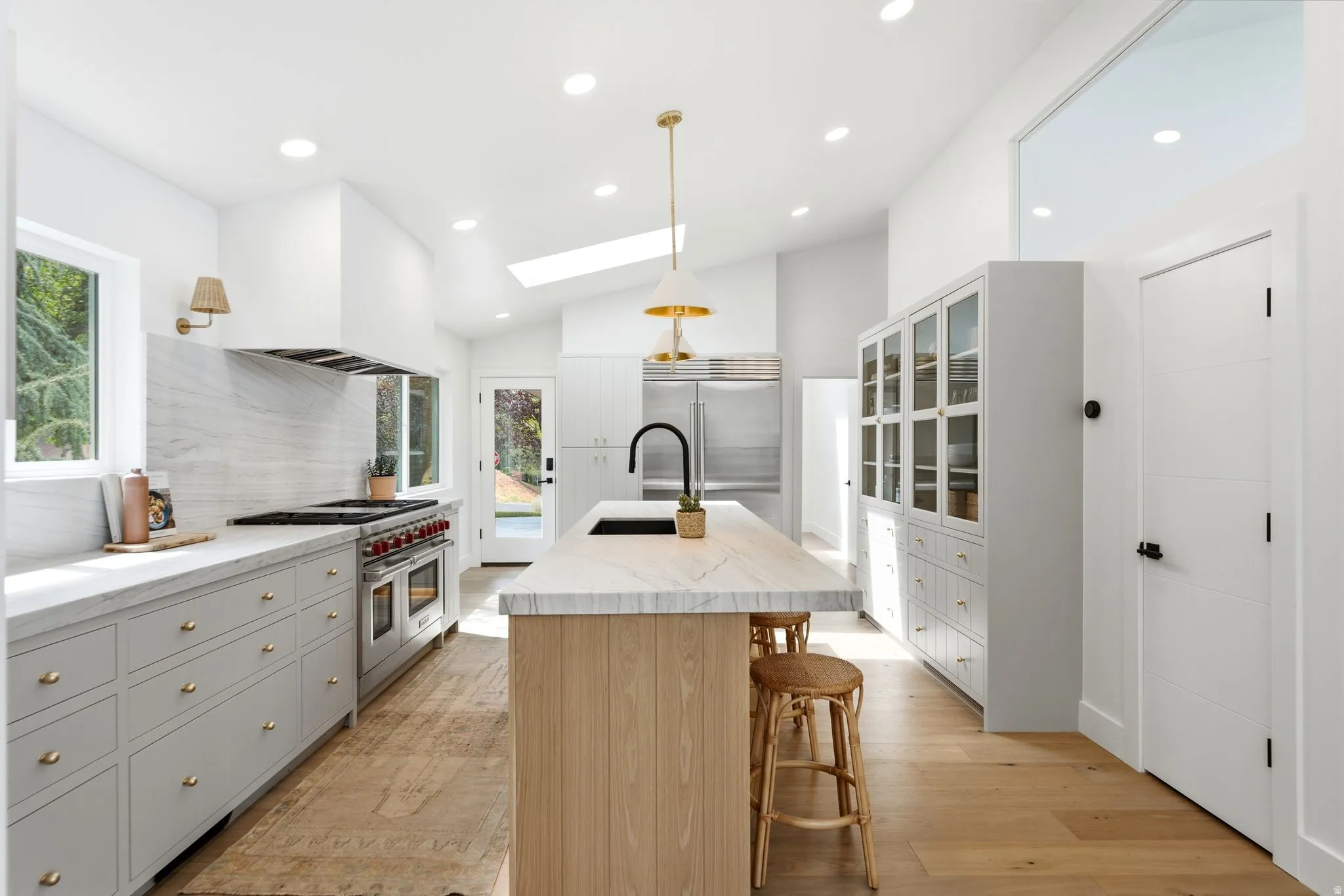 Vaulted ceiling with skylight, wood-finish flooring, central island with sink, extensive cabinetry with glass fronts, and stainless steel appliances