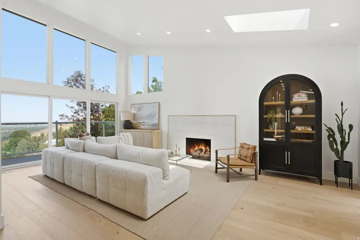 Vaulted ceilings with a large skylight, expansive windows, light wood-finish flooring, and a fireplace with a stone surround