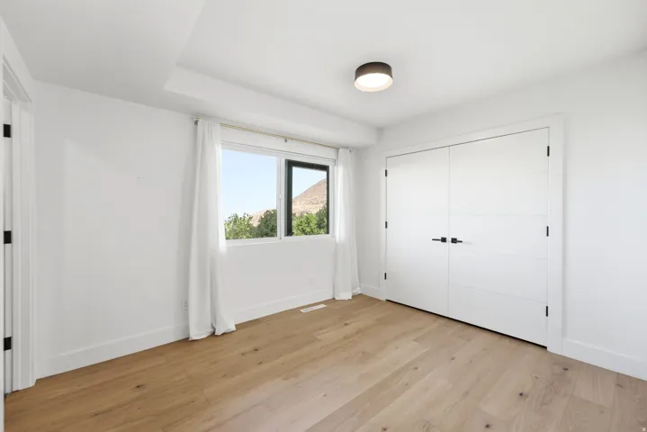 Light wood-finish flooring, white walls, modern flush mount lighting, a large window, and double closet doors with matte black hardware