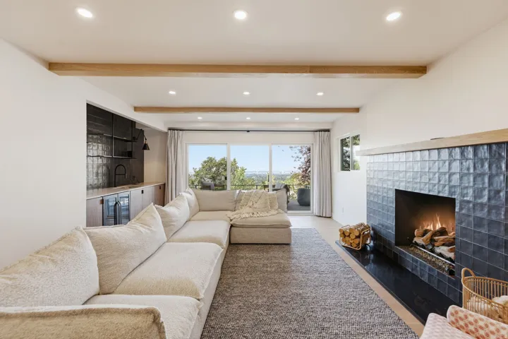 Recessed lighting illuminating the wood-finish ceiling beams, expansive sliding glass doors, a built-in wet bar with dark tile backsplash, and a fireplace with dark tile surround