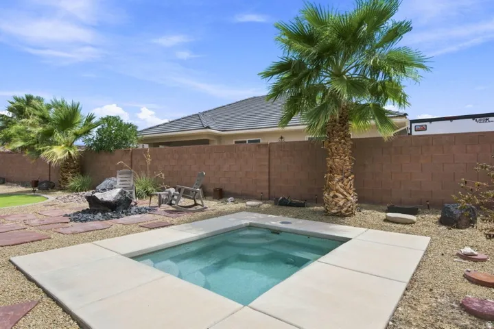 In-ground spa with concrete coping, surrounded by gravel landscaping, mature palm trees, and a block fence