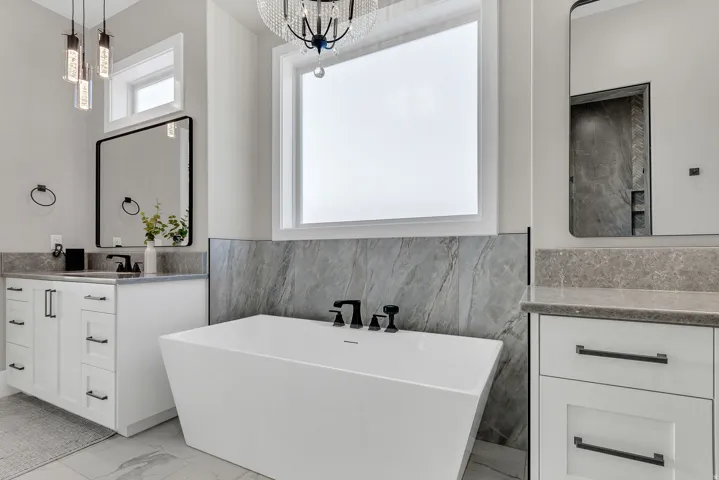Freestanding soaking tub with matte black fixtures, white shaker cabinetry with dark hardware, stone countertops, and full-height stone accent wall