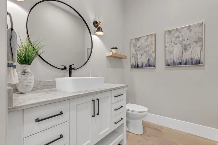 White vanity with black hardware, rectangular vessel sink, round wall mirror, light wood-finish flooring, and a floating shelf