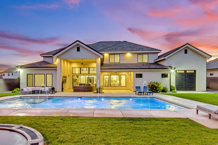 Spacious backyard featuring a rectangular swimming pool with integrated water features, expansive concrete patio, manicured lawn, and a two-story residence with a covered outdoor living area and multiple large windows