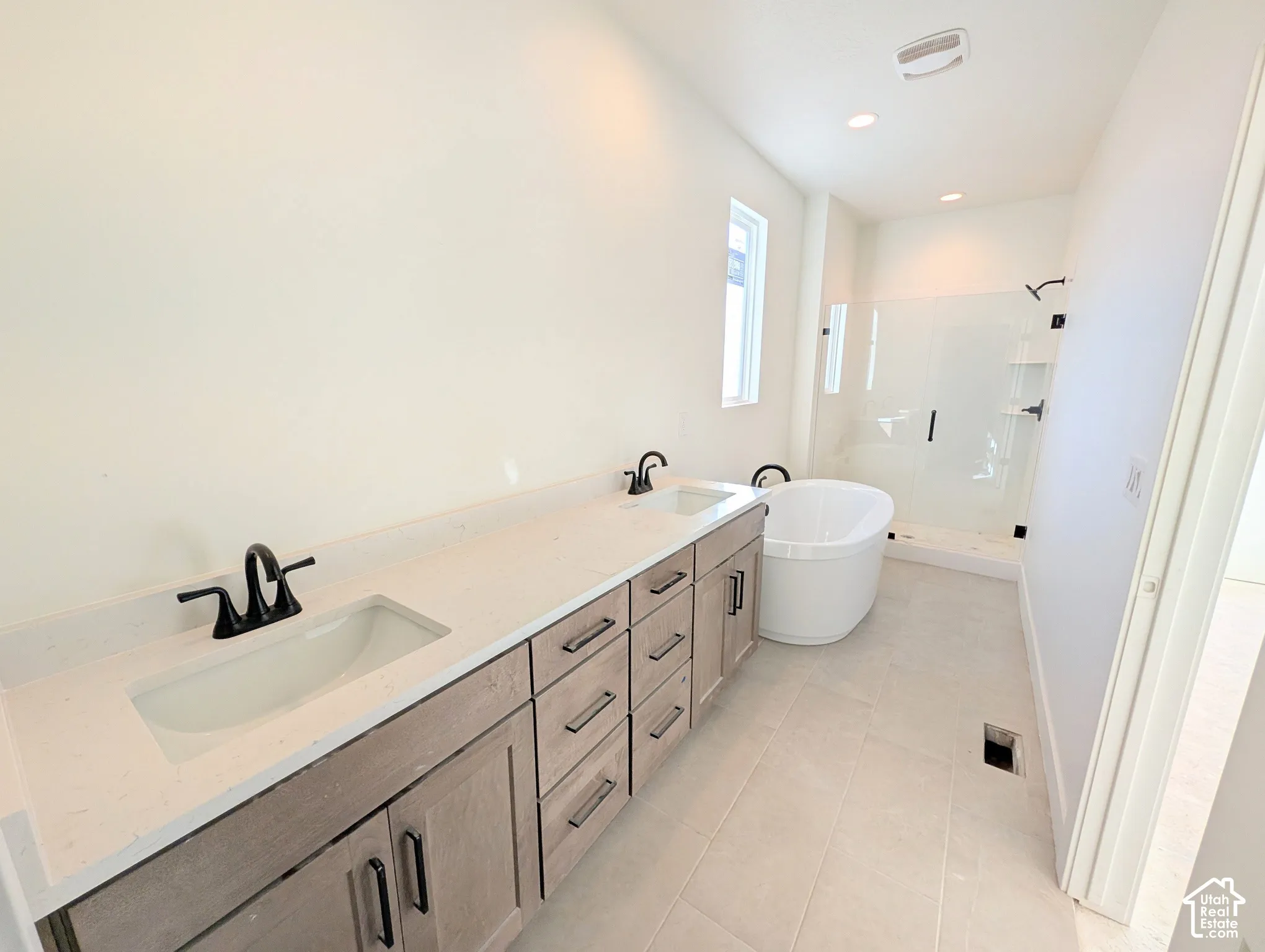 Dual sink vanity with light wood-finish cabinetry, white countertop, matte black fixtures, free-standing tub, and glass-enclosed shower
