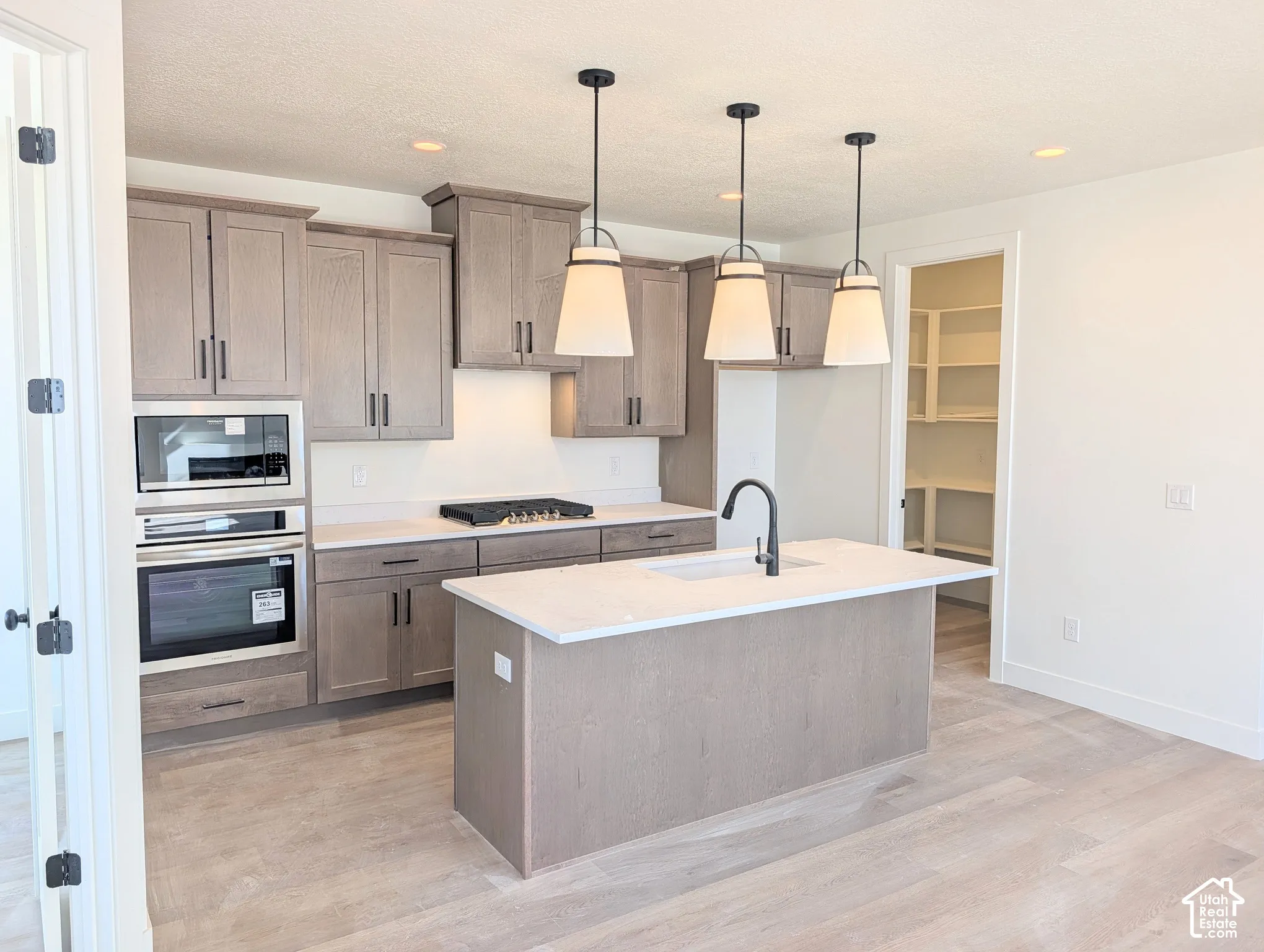 Kitchen featuring wood-finish cabinetry, built-in stainless steel appliances, a center island with a sink, pendant lighting, and a pantry with built-in shelving