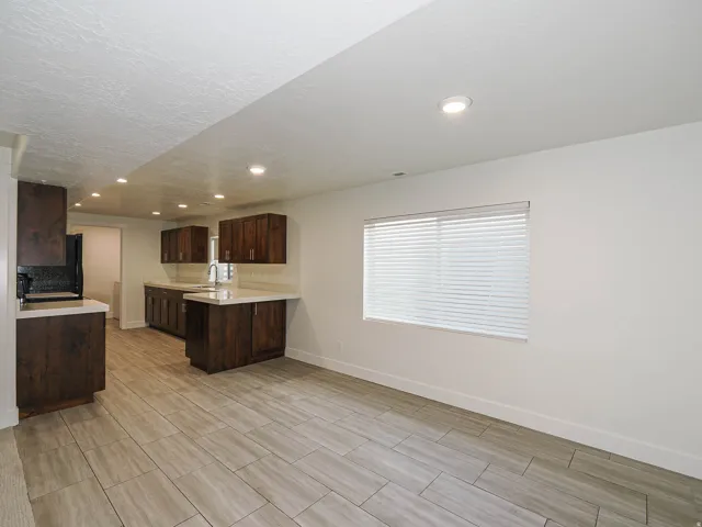 Modern kitchen and dining area featuring dark wood cabinetry, a centered island with seating, stainless steel appliances, recessed ceiling lighting, wood-finish flooring throughout, and a large horizontal window with plantation shutters allowing abundant