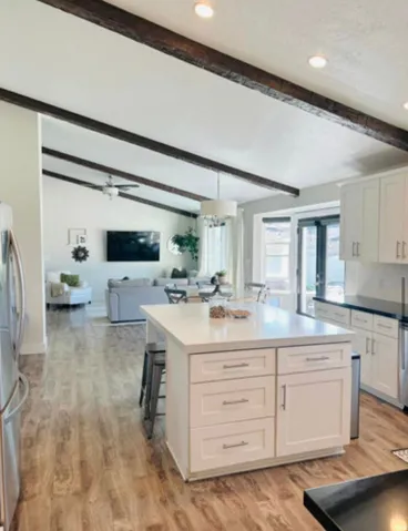 Vaulted ceiling with exposed wood beams, wood-finish flooring, central kitchen island with seating, white shaker cabinetry, and stainless steel appliances