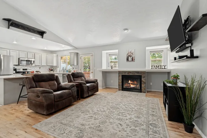 Vaulted ceiling, wood-finish flooring, stone-faced fireplace, white cabinetry with stainless steel appliances, and a kitchen island with seating