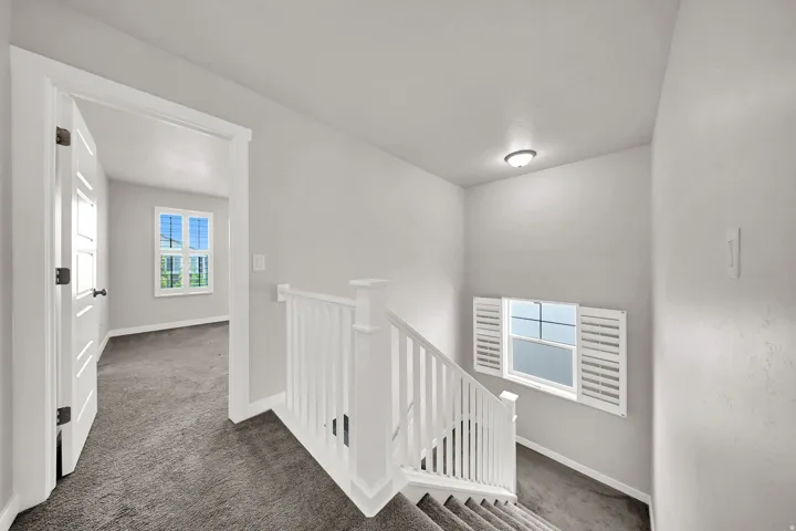 Bright upper-level hallway featuring neutral finishes, recessed ceiling lighting, white painted railing, and louvered window shutters providing natural light and ventilation control