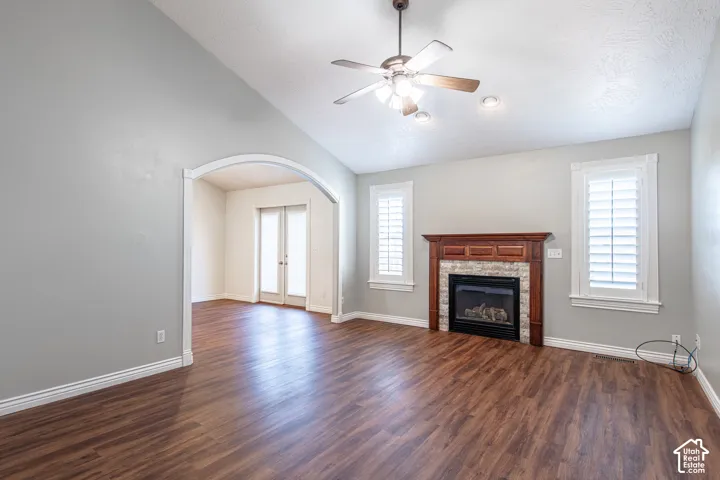 Vaulted ceiling with a ceiling fan, wood-finish flooring, fireplace with a wood mantle and stone surround, and an arched doorway leading to an additional space with French doors