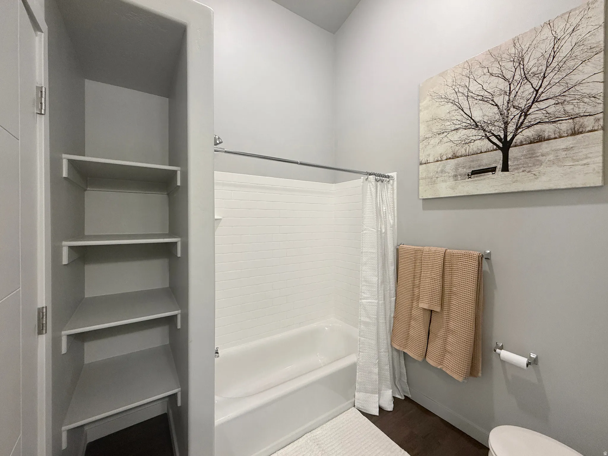 Bathtub with white subway tile surround, built-in shelving, dark wood-finish flooring, and light gray walls