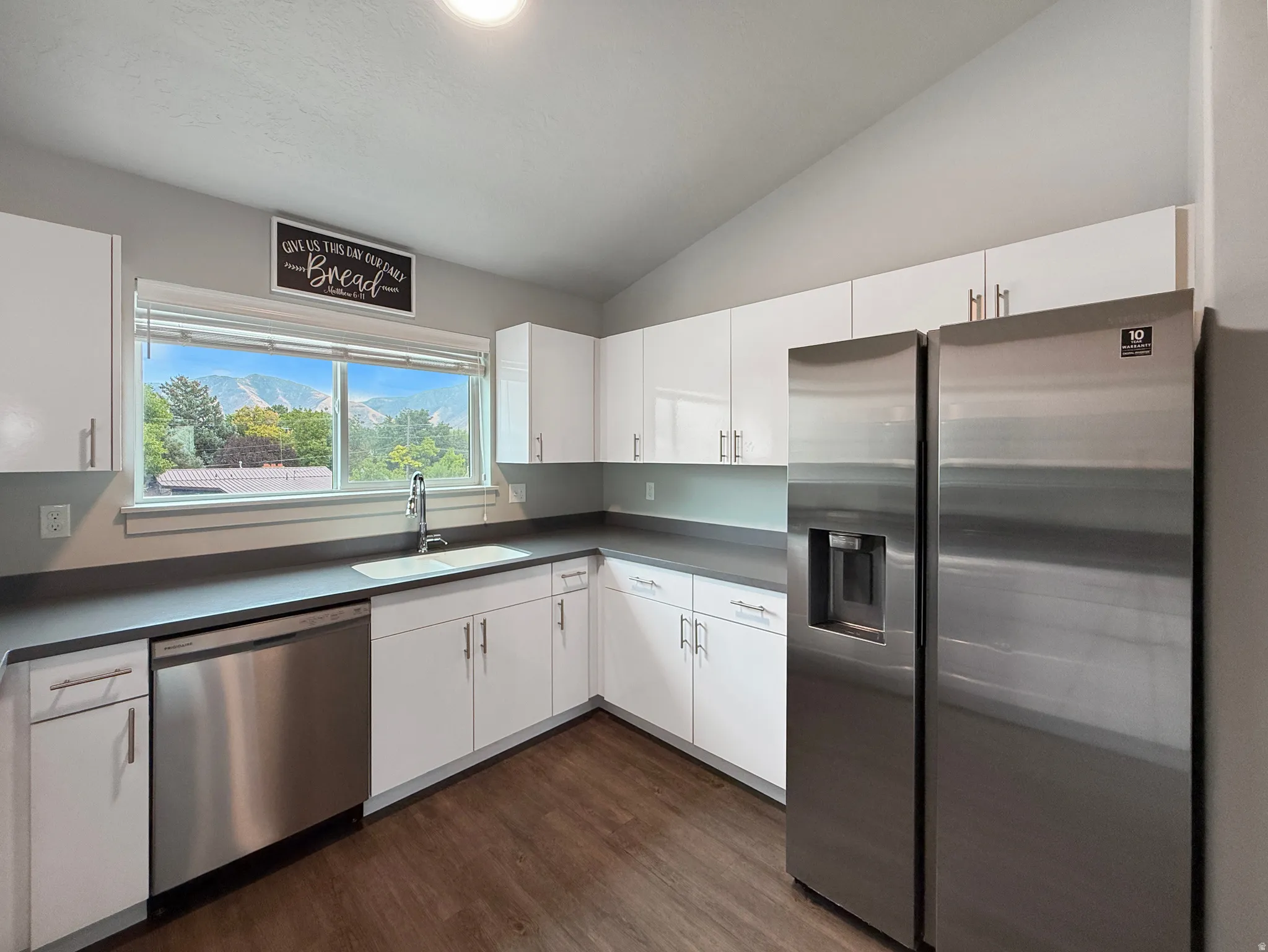 Kitchen featuring white cabinetry, dark countertops, stainless steel appliances, wood-finish flooring, and a vaulted ceiling