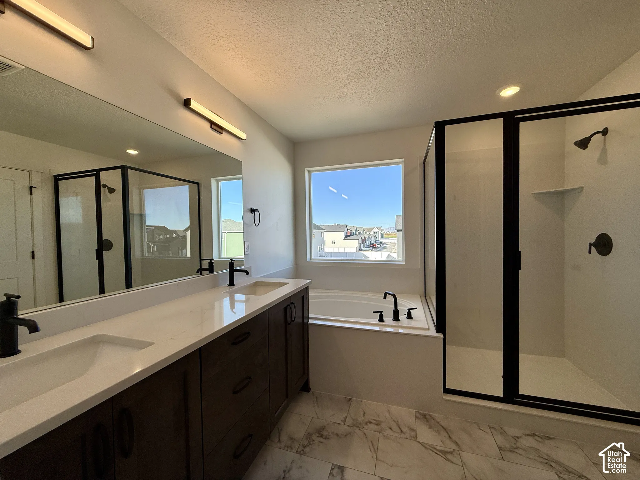 Double vanity with light-toned countertops, large-format tile flooring, soaking tub, glass-enclosed shower with matte black fixtures, and modern vanity lighting