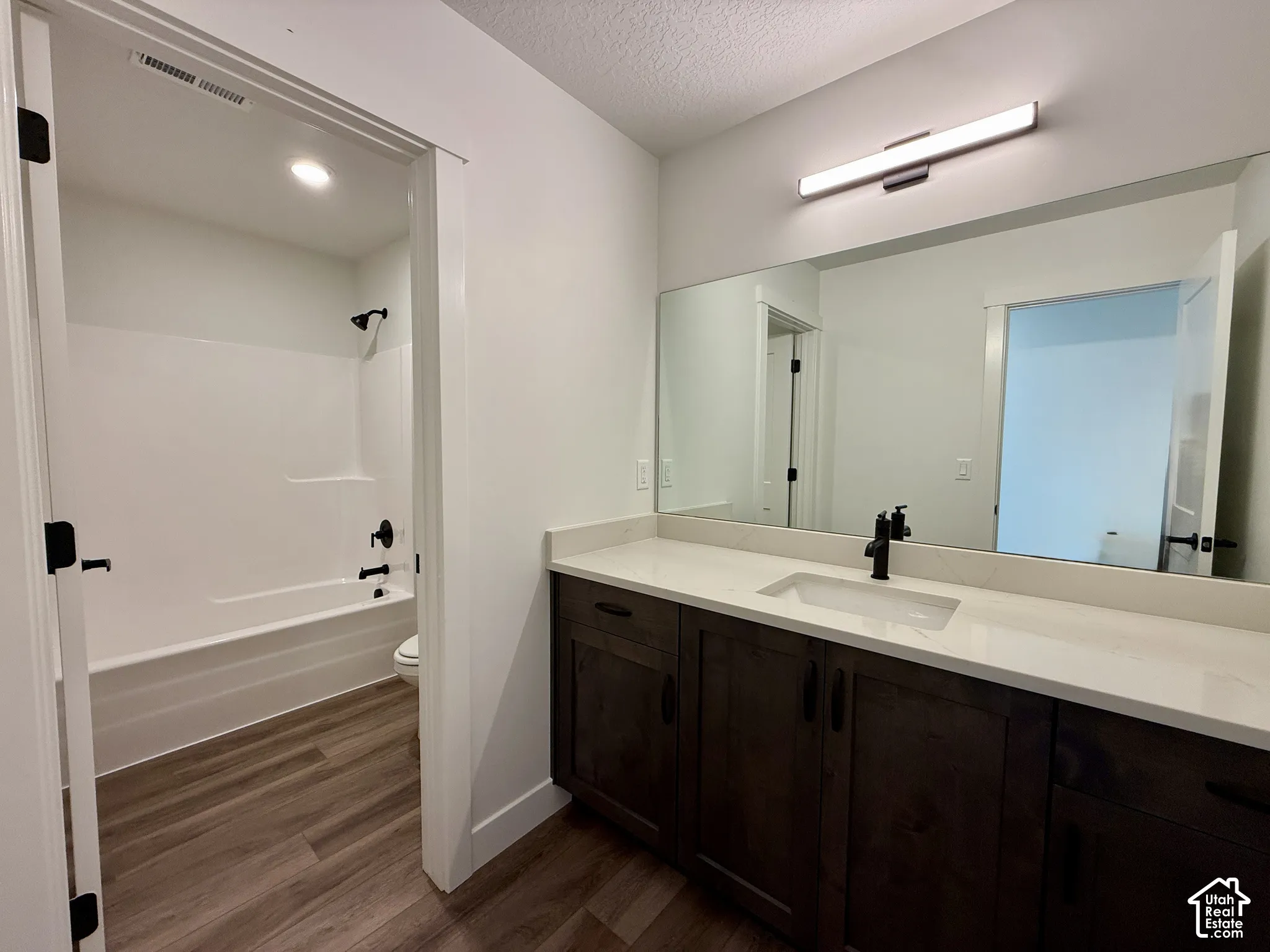 Bathroom featuring dark wood-finish cabinetry, solid-surface countertop with an integrated rectangular basin, matte black fixtures, full-width vanity mirror, and wood-finish flooring