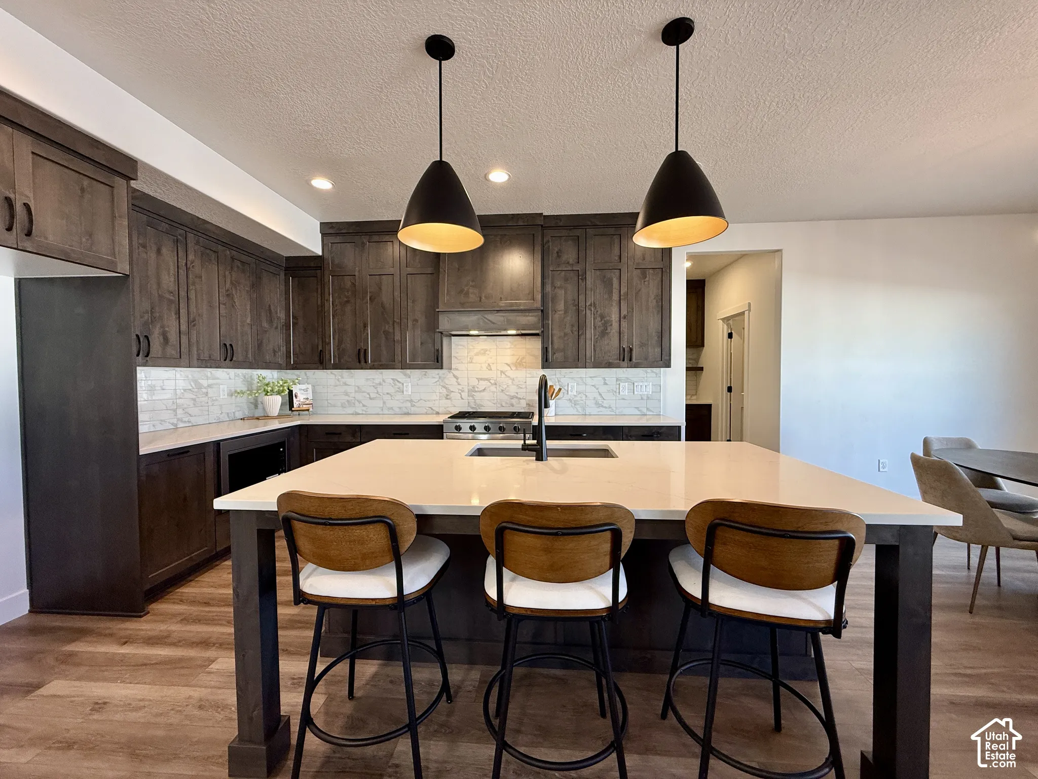 Kitchen featuring custom dark-toned cabinetry, quartz countertops, a central island with an integrated sink, patterned tile backsplash, and matte black pendant lighting