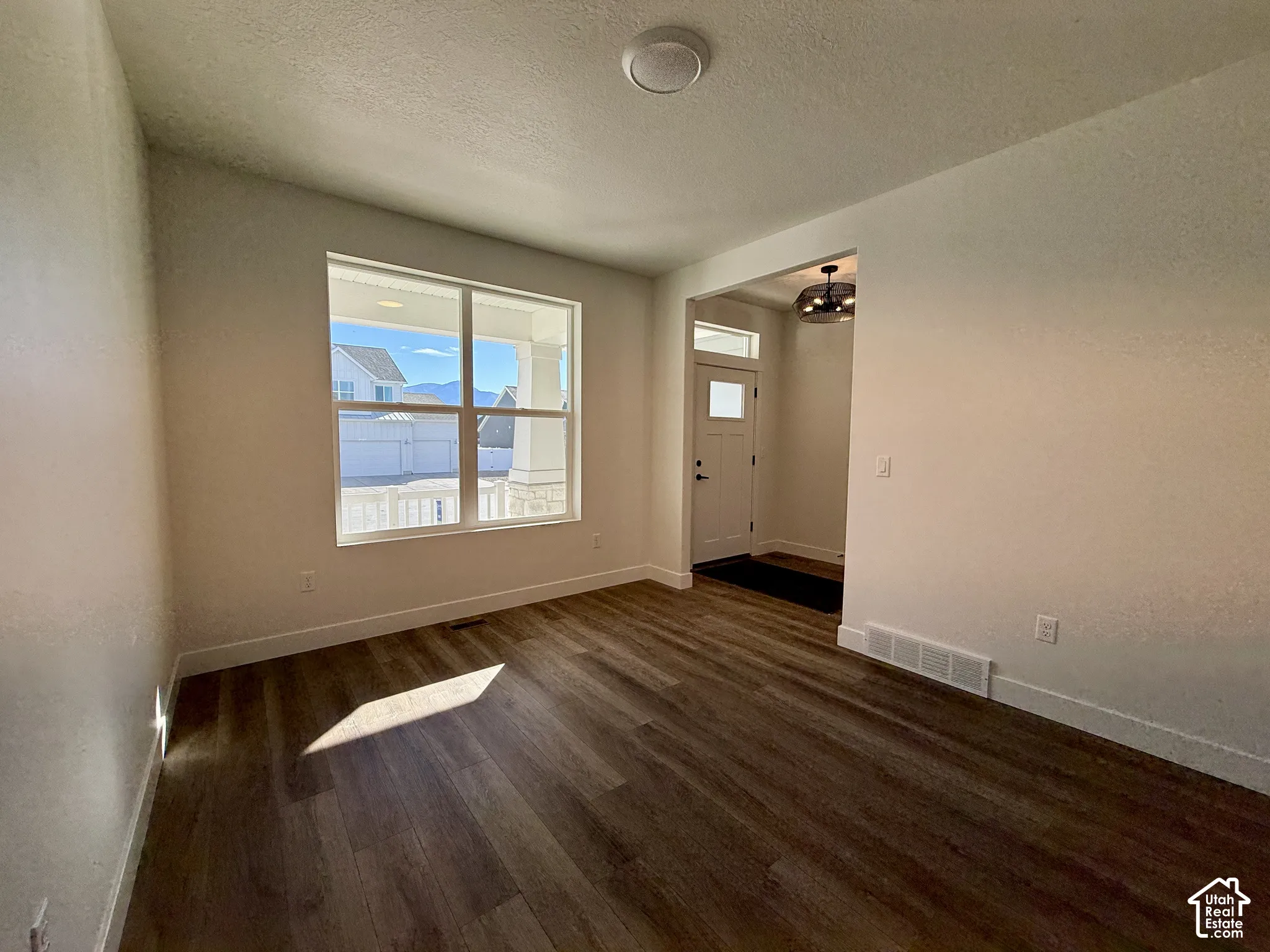 Room featuring wood-finish flooring, a large picture window, and a multi-light flush-mount ceiling fixture, with visibility of an entry door and transom window