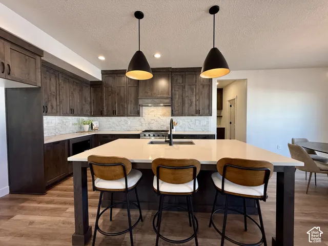 Kitchen featuring custom dark-toned cabinetry, quartz countertops, a central island with an integrated sink, patterned tile backsplash, and matte black pendant lighting