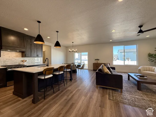 Open-concept living space featuring wood-finish flooring, dark-toned cabinetry, a stone-surround backsplash, a central kitchen island with a neutral countertop, and recessed lighting