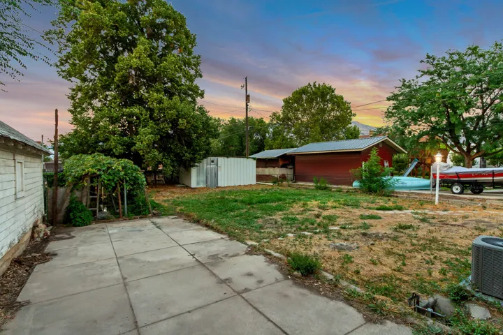 Expansive concrete patio, mature trees providing shade, additional storage shed, and a detached building