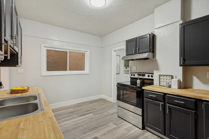 Kitchen featuring wood-finish countertops, stainless steel double basin sink, black cabinetry with brushed nickel hardware, stainless steel range with overhead hood, and light wood-look flooring