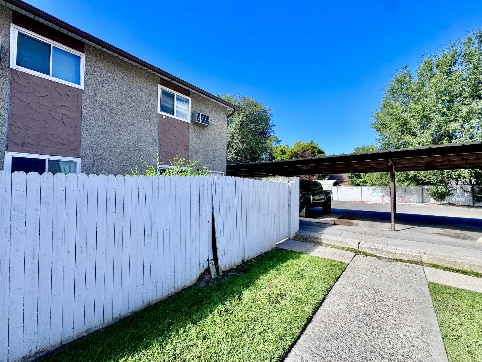 Stucco exterior with brown accents, white picket fence, concrete walkway, green lawn, and covered parking structure
