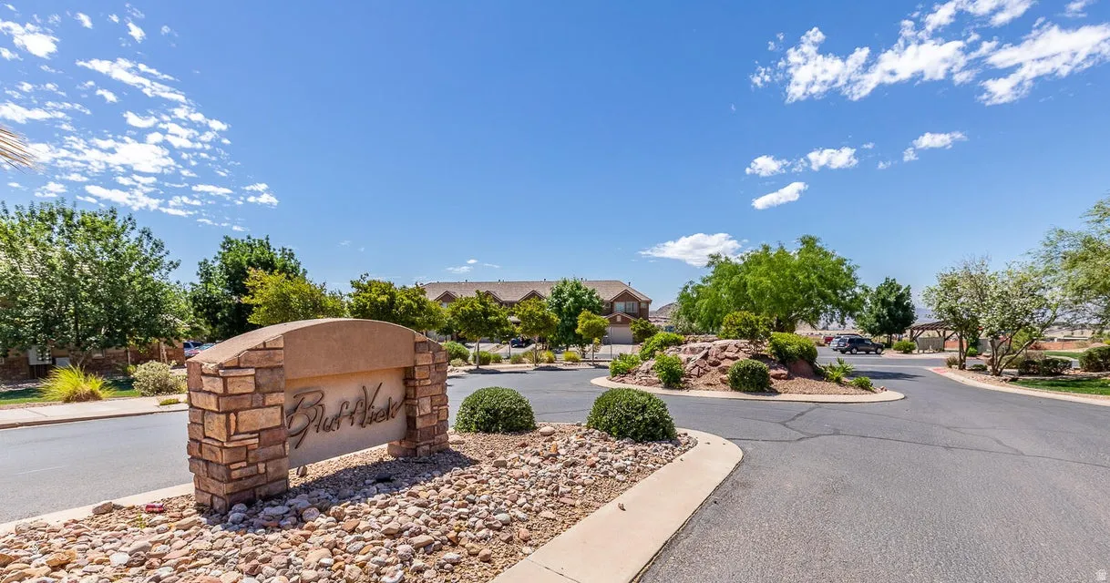 Stone-clad community signage, landscaped median with mature plantings, paved road, and distant residential structures