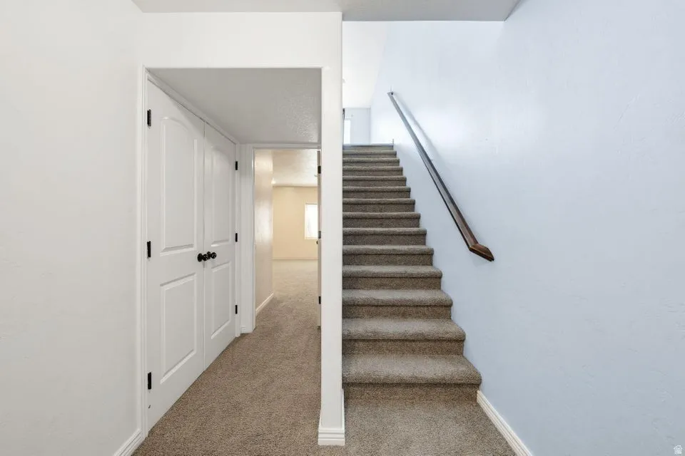 Bright staircase with carpeted treads and metal handrail, opening to a hallway featuring neutral carpeting and clean white walls