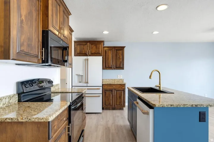 Kitchen featuring wood cabinetry, light-toned granite countertops, a black electric range, a white refrigerator, and a kitchen island with a black sink and brass faucet
