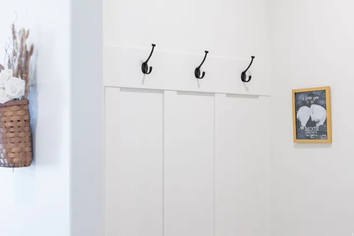 Bright entryway showcasing white shiplap walls, three black wall-mounted hooks for convenient storage and organization, and a framed chalkboard accent piece