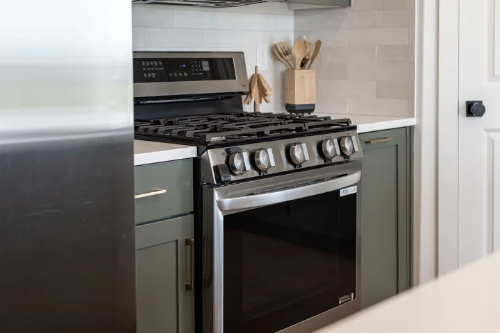 Stainless steel range with a gas cooktop, white countertop, green shaker-style cabinetry with gold-tone hardware, and a light-toned tile backsplash