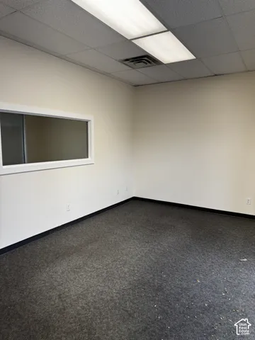 Enclosed office space featuring neutral wall paint, carpeted flooring, a recessed interior window with a white frame, and a suspended drop ceiling with integrated fluorescent lighting