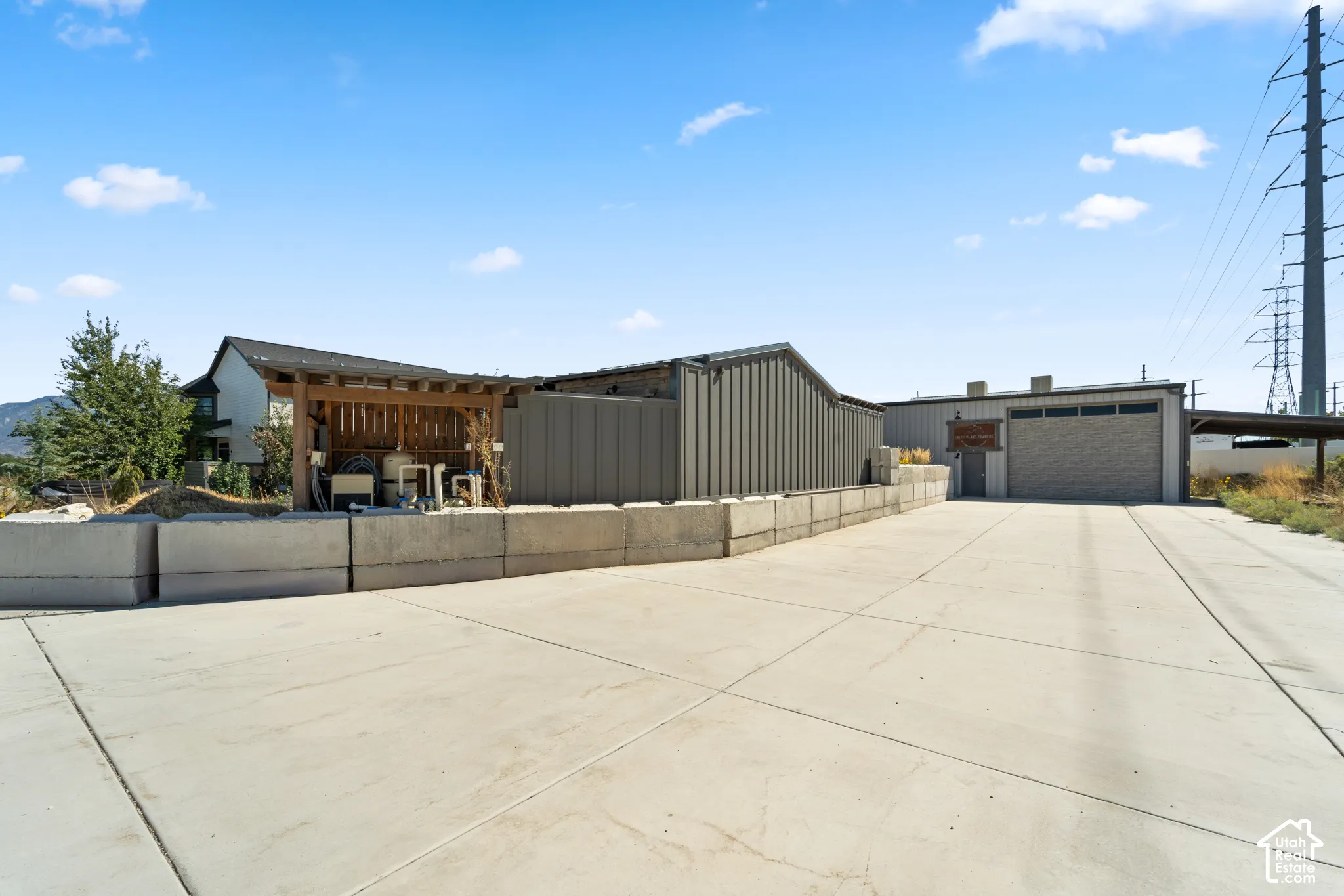 Expansive concrete driveway, a dedicated utility area with a wooden pergola, a large corrugated metal building, and a separate structure with a roll-up garage door