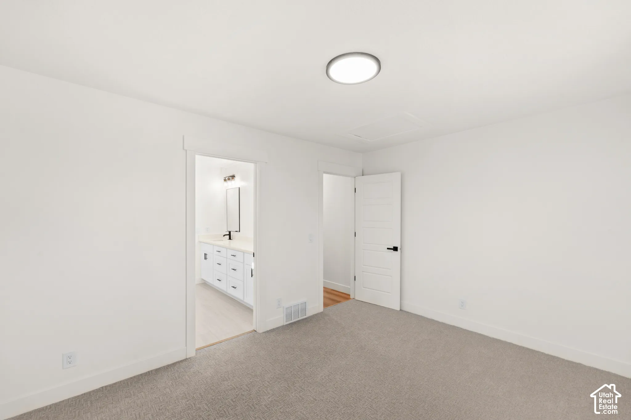 Recessed ceiling light fixture, neutral carpet flooring, white interior doors with black hardware, white walls, and a visible bathroom vanity with a mirror and light fixture
