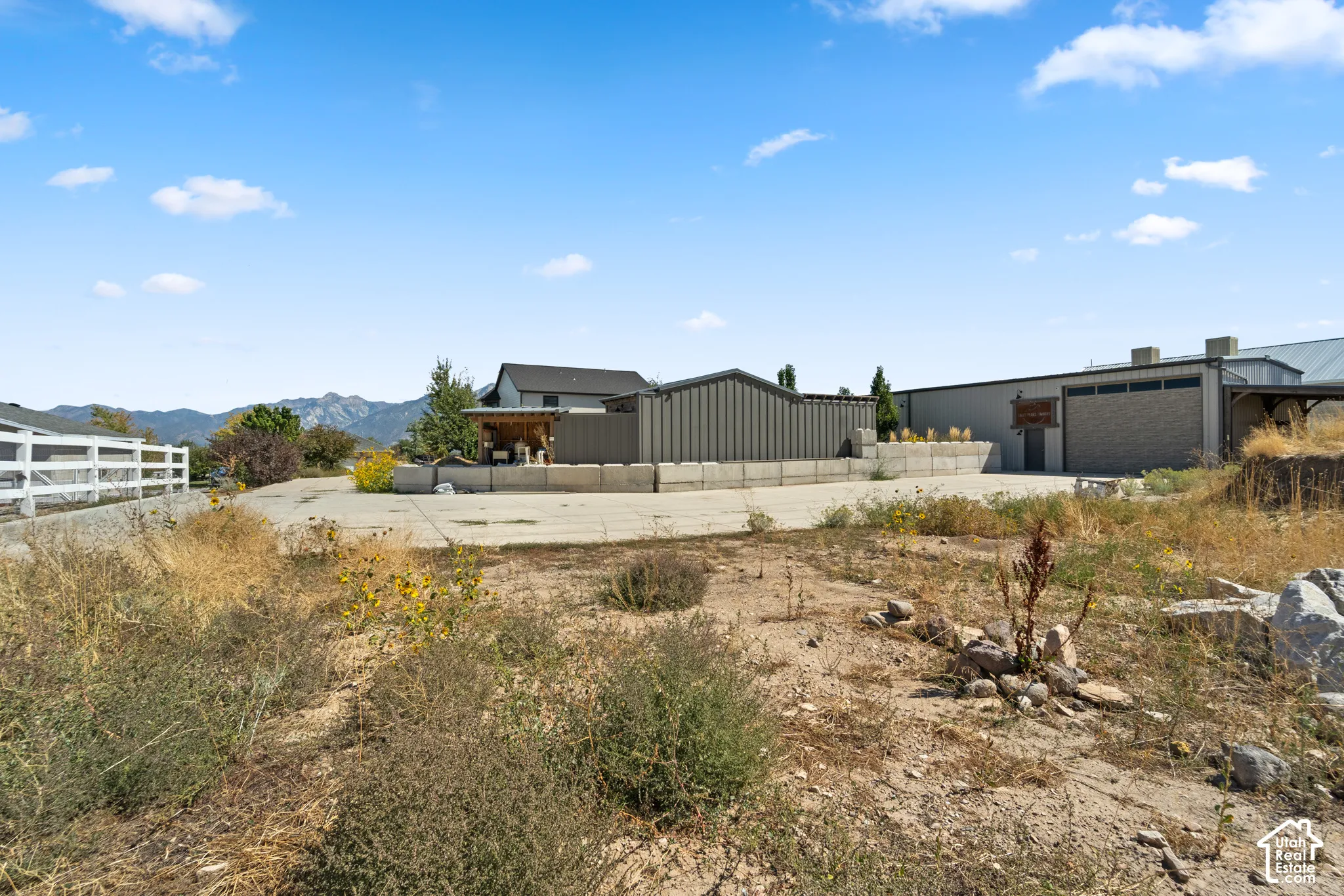 Gated property entrance with a white fence, featuring a large concrete-paved area and mountains in the background