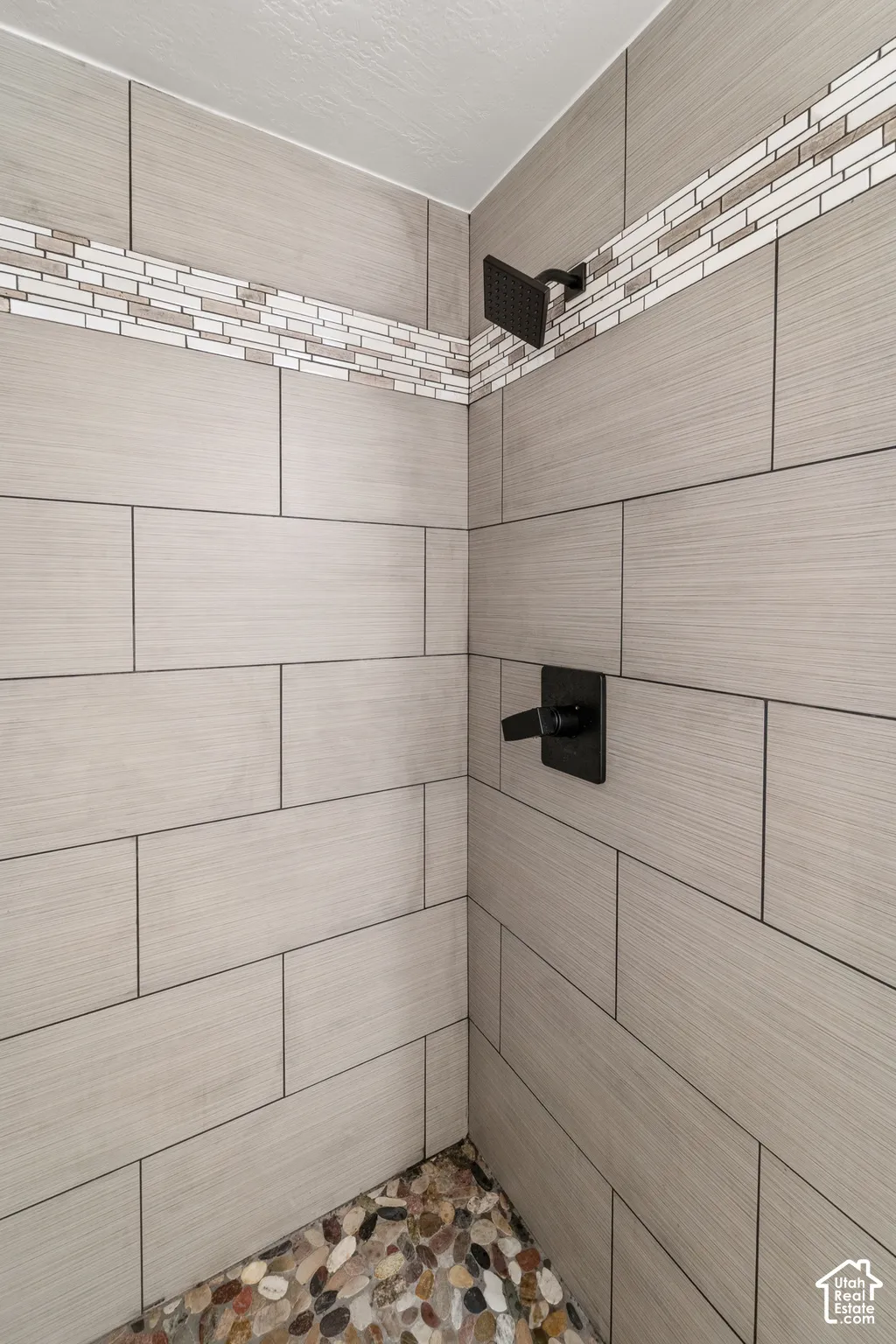 Shower stall featuring large format tile walls, a decorative mosaic tile accent band, a river rock floor, and matte black fixtures