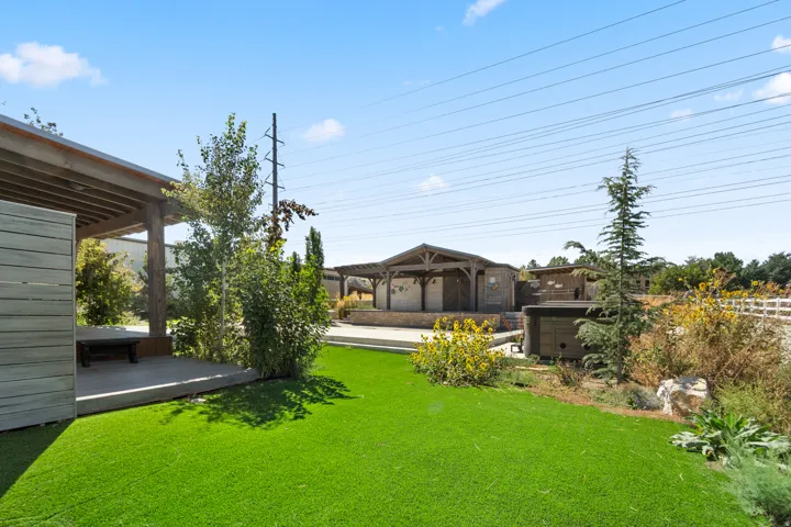 Expansive green space featuring artificial turf, a covered patio with wooden beams, a built-in outdoor structure with a stone facade, and mature landscaping