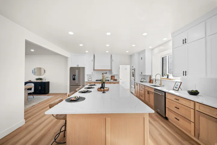 Spacious kitchen featuring an expansive central island with a light-toned countertop, wood-finish flooring, white upper cabinetry, and natural wood-tone lower cabinetry