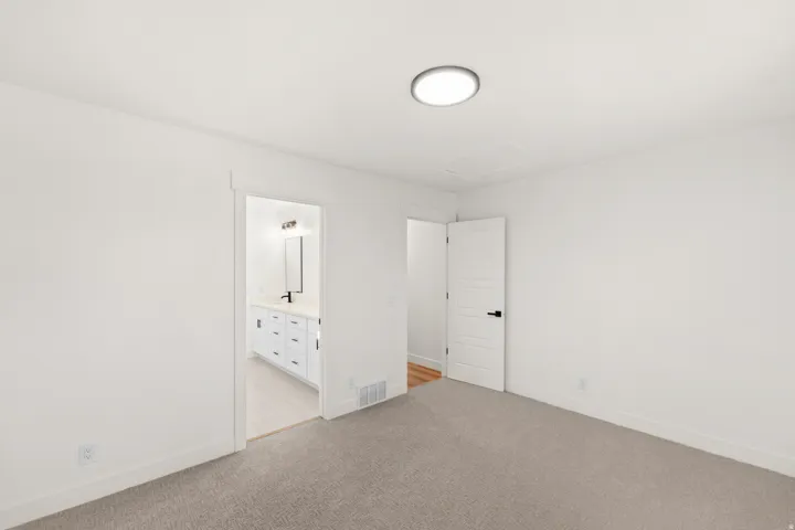 Recessed ceiling light fixture, neutral carpet flooring, white interior doors with black hardware, white walls, and a visible bathroom vanity with a mirror and light fixture