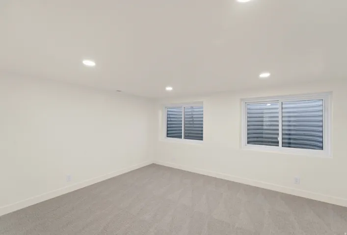 Recessed lighting illuminates this room featuring light-toned walls, grey patterned carpeting, and two windows
