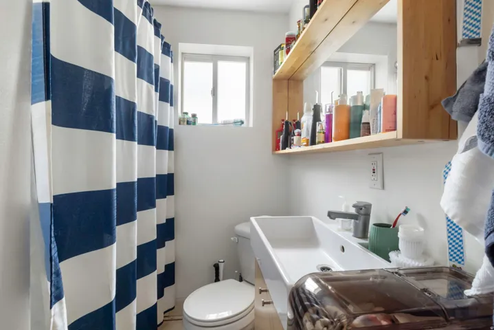 Compact bathroom featuring a window, a wall-mounted vanity with a single basin sink, a toilet, and a wood-finish wall shelf