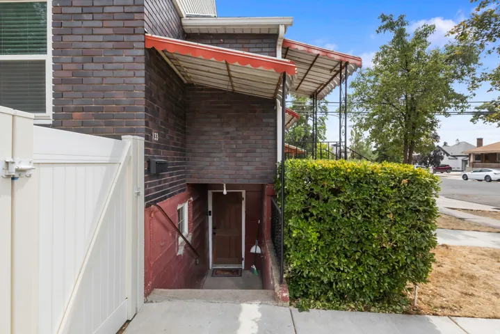 Brick facade, privacy hedge, white vinyl gate, covered entry, and wood-panel door