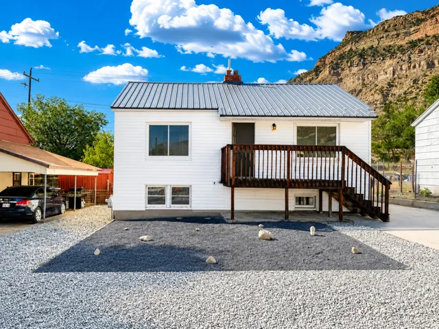 White horizontal siding with a dark metal roof, a wood deck with stairs, a gravel driveway, and a covered parking area