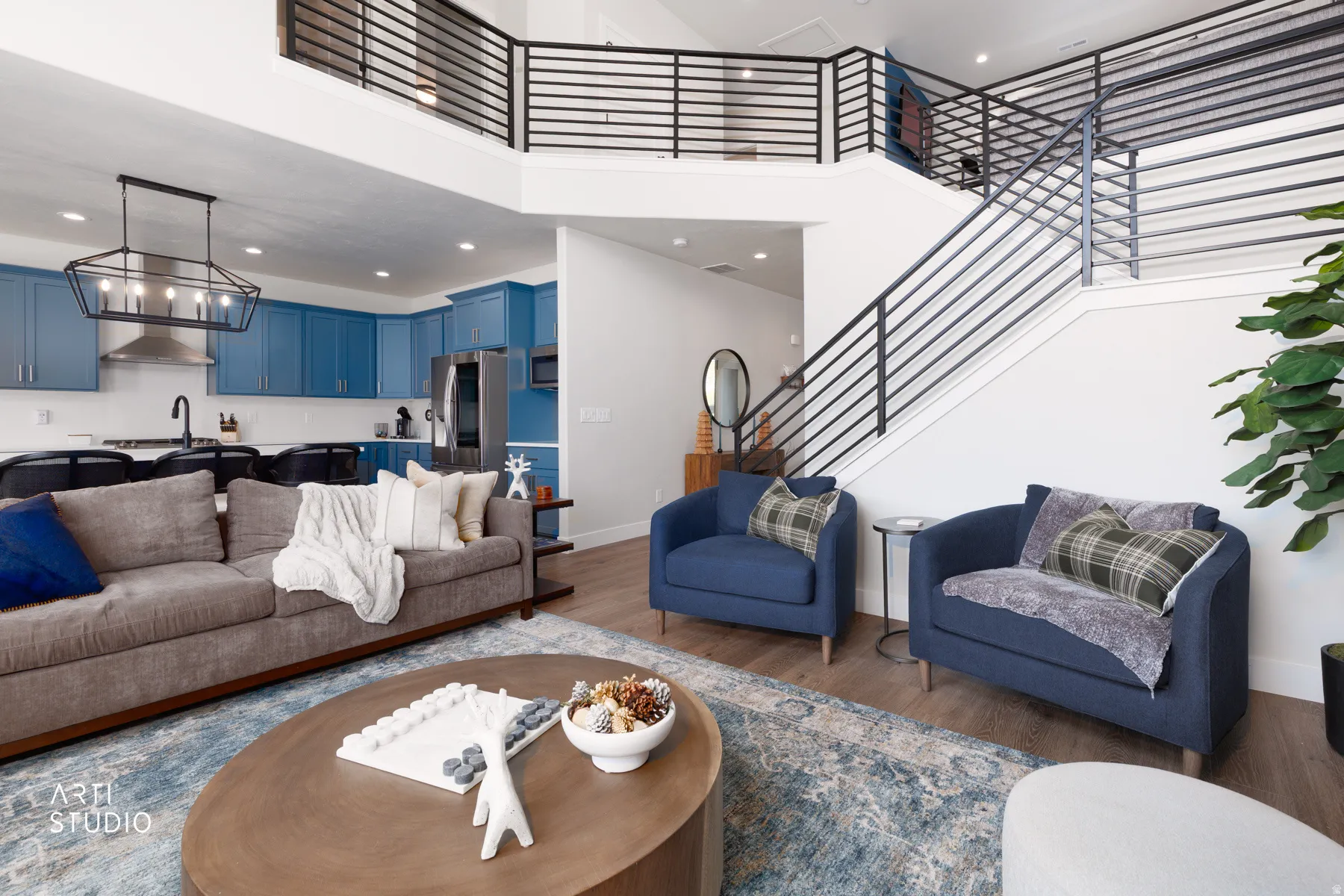 Open-concept living area with wood-finish flooring, a kitchen featuring blue cabinetry and stainless steel appliances, and a staircase with contemporary horizontal railings leading to an upper landing