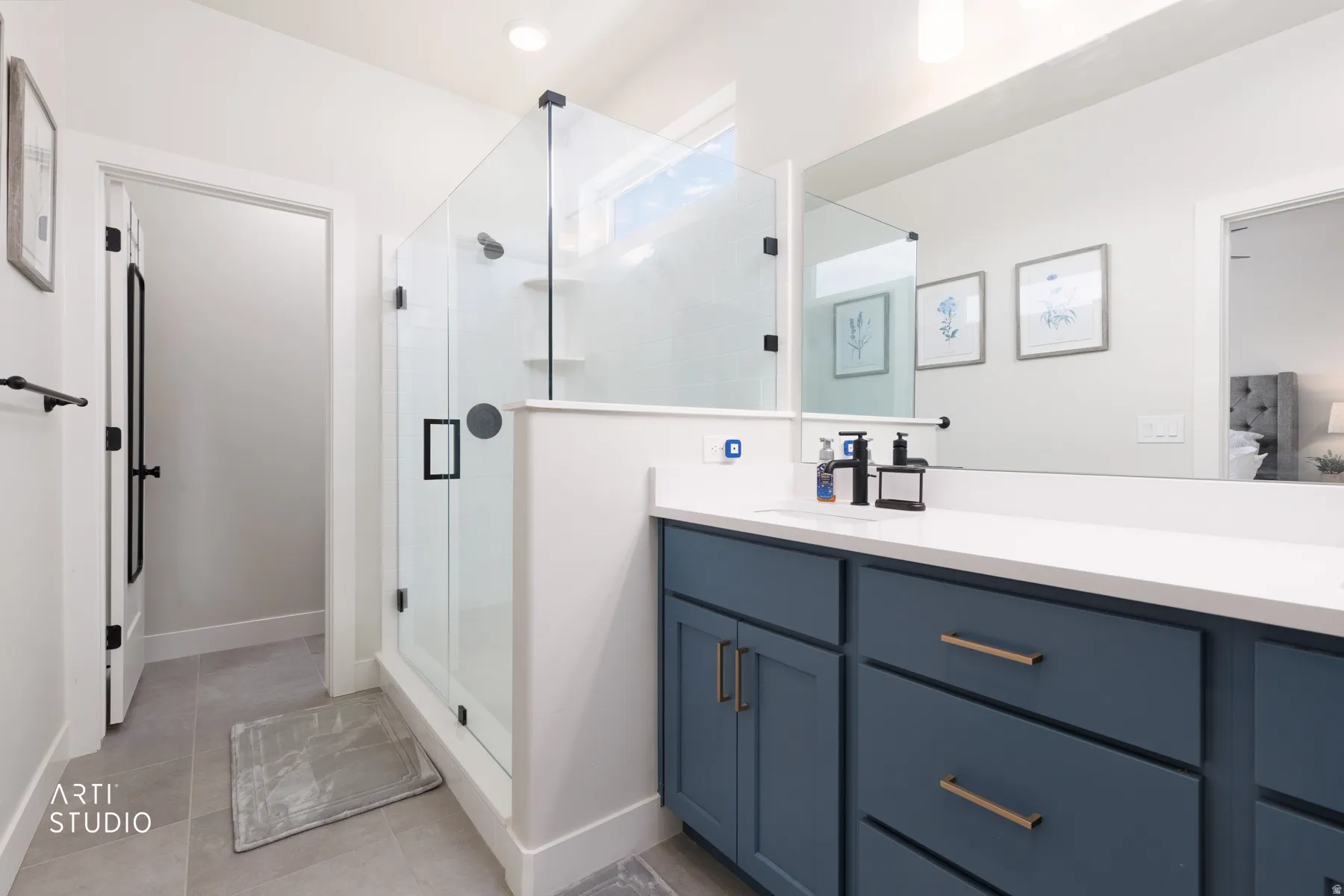 Glass-enclosed shower with black fixtures, long vanity with a solid surface countertop, dual sinks, and blue cabinetry with gold-toned hardware
